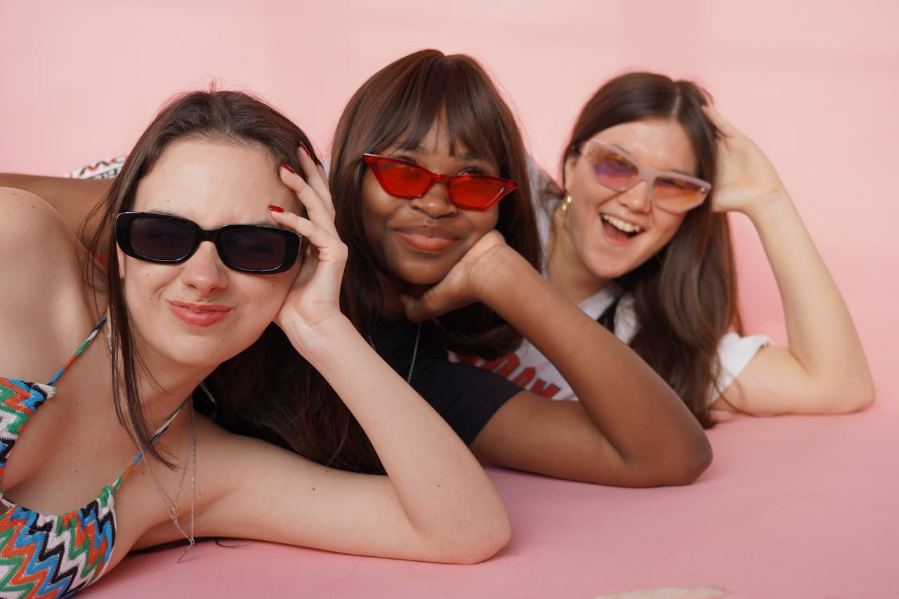 Three joyful women in sunglasses posing playfully in a studio with a pink background.